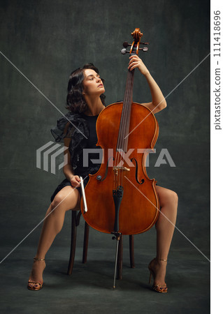 Elegant, beautiful young woman, cellist sitting with wooden cello against dark green vintage background. Acoustic concert Elegant, beautiful young woman, cellist sitting with wooden cello against dark green vintage background. Acoustic concert 111418696