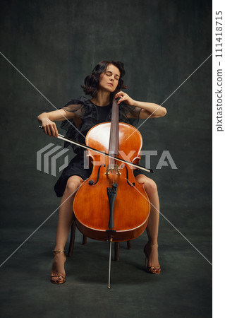 Passionate young woman, musician sitting with eyes closed and playing cello over vintage green background. Being fully into melody Passionate young woman, musician sitting with eyes closed and playing cello over vintage green background. Being fully into melody 111418715