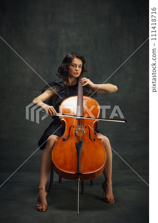Beautiful young woman, cellist sitting on chair with cello, looking at camera against vintage green background. Cultural events with classic music performances. Beautiful young woman, cellist sitting on chair with cello, looking at camera against vintage green background. Cultural events with classic music performances. 111418716