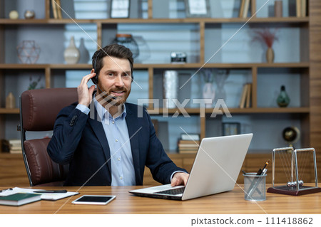 Smiling adult male in business attire multitasking in a home office setup with laptop, headset, and smartphone. 111418862