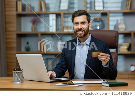 Confident mature businessman in suit with credit card, using laptop at messy home office desk, symbolizing remote work. Confident mature businessman in suit with credit card, using laptop at messy home office desk, symbolizing remote work. 111418929