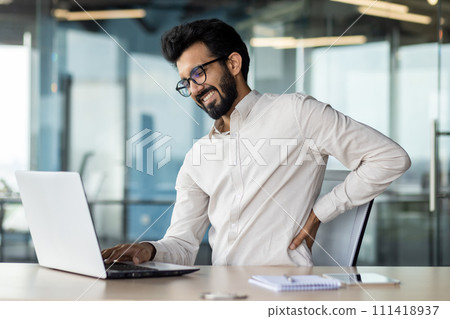 Indian man in glasses feeling back pain while working overtime at his desk with a laptop in a modern office setting. 111418937