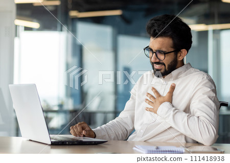 Indian man at office desk suffering from illness or stress while working on laptop, conveying a sense of corporate burnout. 111418938