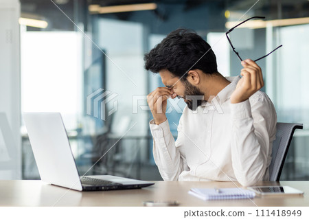 Stressed Indian man at an office desk feeling exhausted and overtired from work, holding his glasses and massaging his nose bridge. 111418949