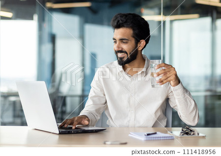 A white-collar worker with a beard is multitasking, drinking water and using a laptop computer on a desk. A white-collar worker with a beard is multitasking, drinking water and using a laptop computer on a desk. 111418956