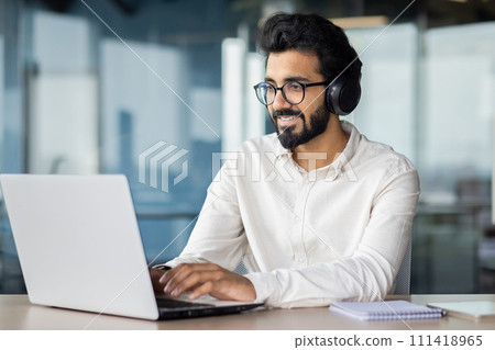 A white-collar worker with a beard is using a laptop computer while sitting at a desk with headphones. A white-collar worker with a beard is using a laptop computer while sitting at a desk with headphones. 111418965