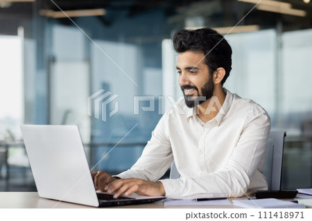 A white-collar worker is using a laptop on a desk, surrounded by office equipment. His beard adds to his professional appearance. A white-collar worker is using a laptop on a desk, surrounded by office equipment. His beard adds to his professional appearance. 111418971