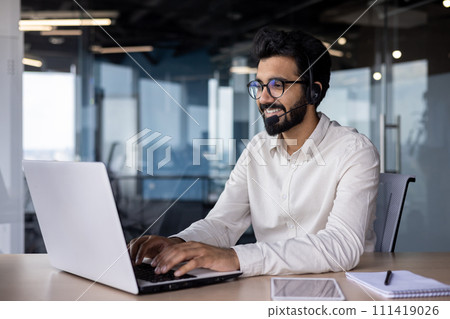 Smiling and confident young Indian man in headset sitting at desk in office and working on laptop. 111419026