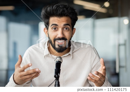 Indian businessman with a beard engaging an audience, speaking into a microphone at a modern office. 111420175