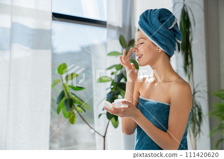 Cute caucasian woman with towel on wet hair, applying face cream on nose after showering. Side view of happy female with closed eyes, using face cream in bathroom. Concept of skincare cosmetic. 111420620