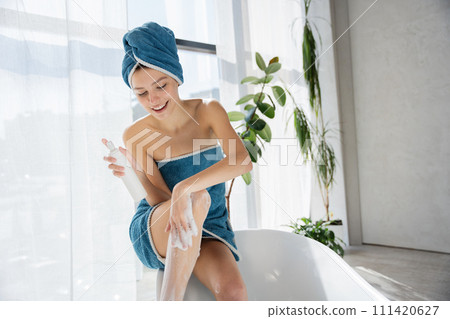 Smiling young woman with towel over head, resting in bathtub, shaving leg at home. Front view of pretty caucasian lady using razor, while sitting in bathtub, against sunlit window. Body care concept. Smiling young woman with towel over head, resting in bathtub, shaving leg at home. Front view of pretty caucasian lady using razor, while sitting in bathtub, against sunlit window. Body care concept. 111420627