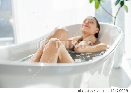 Smiling blonde woman with closed eyes, relaxing in bathtub in spacious bathroom. Front view of silhouette of blurred female with happy face, soaking in bubble bath with closed eyes. Concept of relax. 111420629
