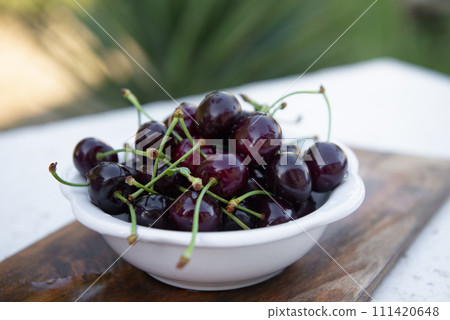 Close up of white plate, board of wet cherries, merries laying on wooden board, table outdoors. Concept of summer. Close up of white plate, board of wet cherries, merries laying on wooden board, table outdoors. Concept of summer. 111420648