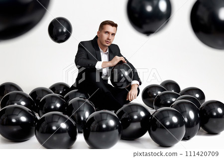 elegant caucasian man in suit tuxedo sitting on studio floor with a lot of black air balloons 111420709