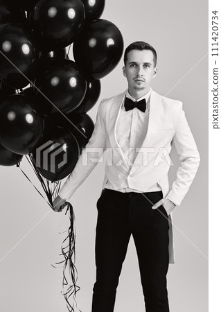 elegant caucasian man in white suit tuxedo with bunch of black air balloons on white background. 111420734