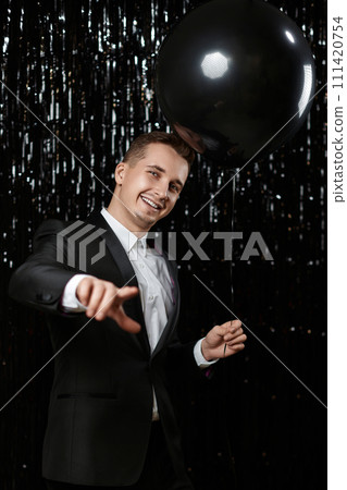 Happy man in black jacket with black air balloons points at camera choosing you on glitter background. 111420754