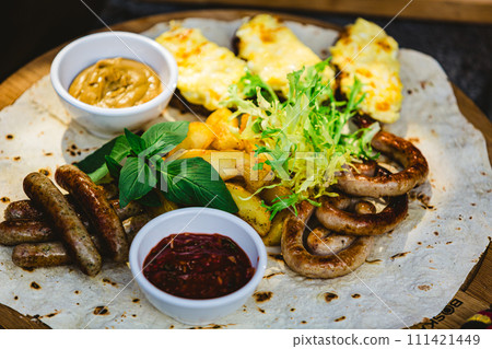 Grilled sausages with potato wedges and bread with cheese and sauces close-up on a wooden tray. 111421449