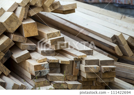 Close-up of piled stack of natural brown uneven rough wooden boards lit by bright sun. Industrial timber for carpentry, building, repairing and furniture, lumber material for construction. Close-up of piled stack of natural brown uneven rough wooden boards lit by bright sun. Industrial timber for carpentry, building, repairing and furniture, lumber material for construction. 111422694