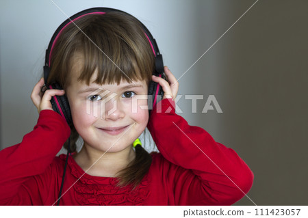Picture of happy pretty child little girl with big headphones at home. Joyful child girl listening to music on gray blurred background. Home, technology and music concept. Picture of happy pretty child little girl with big headphones at home. Joyful child girl listening to music on gray blurred background. Home, technology and music concept. 111423057
