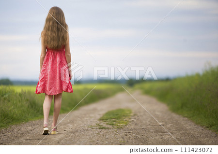 Back view of young romantic slim woman in red dress with long hair walking by ground road along green field on sunny summer day on blue sky copy space background. 111423072