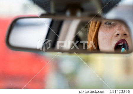 Closeup of a young redhead woman driver correcting her makeup with dark red lipstick looking in car rearview mirror behind steering wheel of a vehicle. Closeup of a young redhead woman driver correcting her makeup with dark red lipstick looking in car rearview mirror behind steering wheel of a vehicle. 111423206