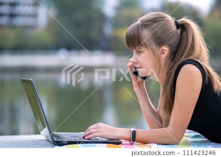 Young female office employee working behind laptop computer while talking on cellphone lying down in summer park outdoors. Remote work concept. 111423246