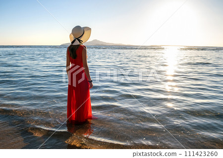 Young woman wearing long red dress and straw hat standing in sea water at the beach enjoying view of rising sun in early summer morning. 111423260