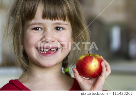 A cute little curly toothless girl smiles and holds a red apple. Portrait of a happy baby eating a red apple. The child loses milk teeth. Healthy food nutrition. 111423262