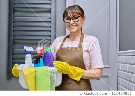 Middle-aged woman in gloves apron with basin of detergents in bathroom Middle-aged woman in gloves apron with basin of detergents in bathroom 111424991