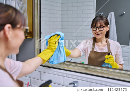 Middle-aged woman in an apron cleaning mirror in bathroom 111425573