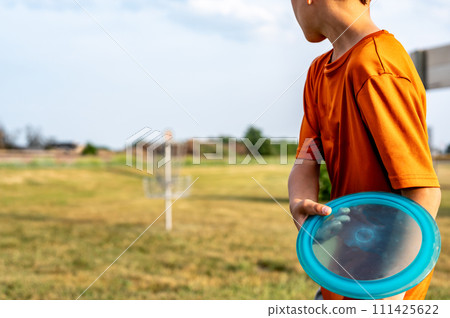 Selective focus fairway grass with a defocused young child holding a putter disc and golf goal in the distance.  111425622
