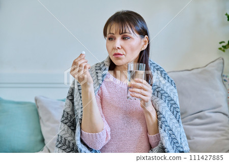 Sick mature woman taking pill with glass of water, sitting under blanket at home Sick mature woman taking pill with glass of water, sitting under blanket at home 111427885