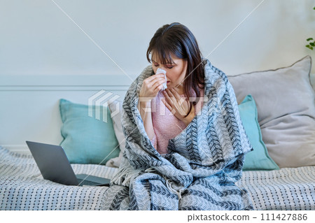 Sick mature woman with blanket, sneezing coughing blowing nose in napkin Sick mature woman with blanket, sneezing coughing blowing nose in napkin 111427886