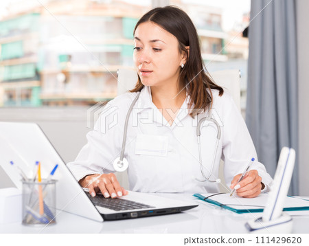 Attentively female doctor working at laptop and writing papers 111429620