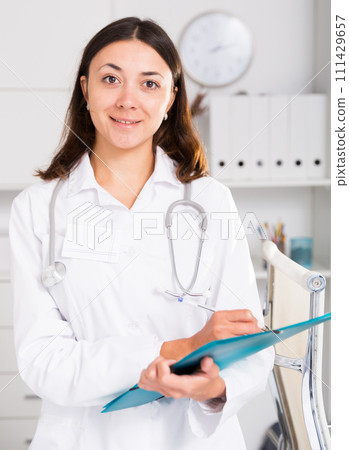 Young positive female medic in white uniform holding clipboard Young positive female medic in white uniform holding clipboard 111429657