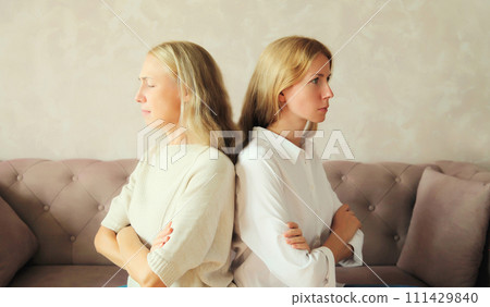 Upset offended middle aged mother or sister and adult daughter together sitting on couch at home 111429840