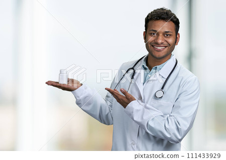 Handsome smiling doctor with stethoscope presenting pills on blur clinic interior background. People, medicine and pharmacology concept. 111433929