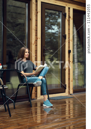 Cute woman in casual clothing sits reading on a wet terrace of a log cabin after the rain, enjoying fresh air Cute woman in casual clothing sits reading on a wet terrace of a log cabin after the rain, enjoying fresh air 111441055