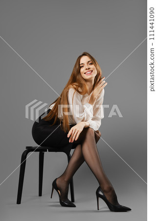 Smiling woman in pencil skirt and blouse leaning forward while sitting on a chair 111441090
