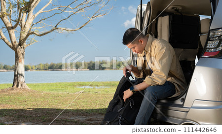 Man traveler sitting in the open trunk of minivan near near a scenic lake under the blue sky 111442646
