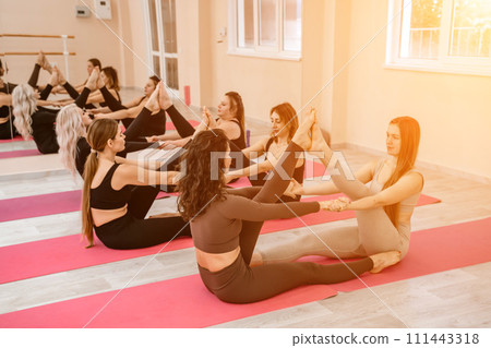 A group of six athletic women doing pilates or yoga on pink mats in front of a window in a beige loft studio interior. Teamwork, good mood and healthy lifestyle concept. A group of six athletic women doing pilates or yoga on pink mats in front of a window in a beige loft studio interior. Teamwork, good mood and healthy lifestyle concept. 111443318