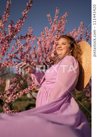 Woman blooming peach orchard. Against the backdrop of a picturesque peach orchard, a woman in a long pink dress and hat enjoys a peaceful walk in the park, surrounded by the beauty of nature. Woman blooming peach orchard. Against the backdrop of a picturesque peach orchard, a woman in a long pink dress and hat enjoys a peaceful walk in the park, surrounded by the beauty of nature. 111443420