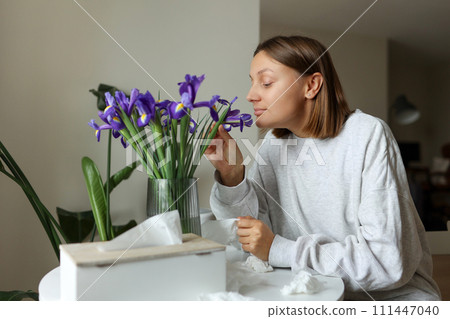 Seasonal allergy free. Young woman sniffs of iris flowers, enjoys of smell without of runny nose, itching or cough seasonal symptoms at cozy home. Girl sits by the table. Used paper tissues on table 111447040