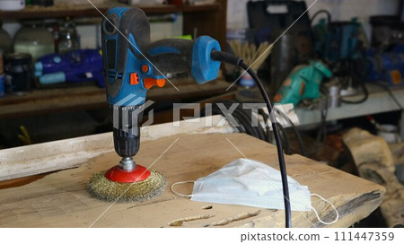 a drill with an attachment made of metal bristles standing on a wooden board in a craftsman's workshop, an apparatus for working with wood and a dust mask as a carpenter's working tools on a workbench 111447359