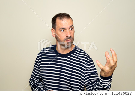 Bearded hispanic man in his 40s wearing a sweater looking extremely angry showing his claw hand while having an aggressive and defiant expression, isolated over beige background. 111450028