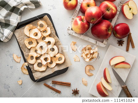 Sweet dried apple rings on baking tray with knife and kitchen towel and red ripe apples. 111451024