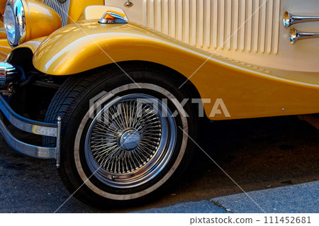 A close-up of a yellow vintage cars front side, showcasing the detailed wire-spoke wheel, chrome bumper, and sleek body design. A close-up of a yellow vintage cars front side, showcasing the detailed wire-spoke wheel, chrome bumper, and sleek body design. 111452681