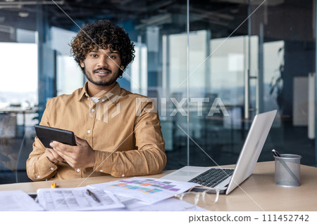 Professional Indian businessman working attentively on a digital tablet, laptop, and documents spread out on his office desk. 111452742