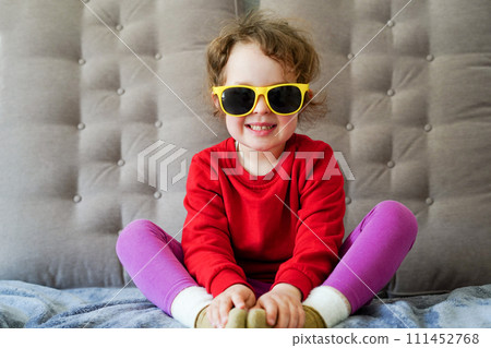 Pretty little girl in red cloth and yellow sunglasses smiling and sitting on the sofa at home 111452768