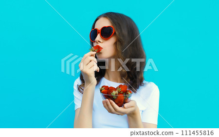 Portrait of woman with handful of fresh strawberries blowing lips in red heart shaped sunglasses 111455816